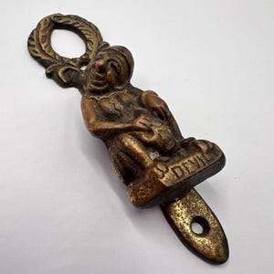 Op de afbeelding: Antieke messing flesopener met een zittende duivelsfiguur. De opener heeft een lus aan de bovenkant en het woord "DEVIL" is op de basis gegraveerd. Het metaal heeft een aangetaste, verouderde uitstraling.