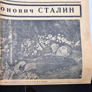 May include: A black and white image of a newspaper page. The headline reads "ОНОВИЧ СТАЛИН". The image shows a man lying down, surrounded by flowers. The text at the bottom is in a foreign language.