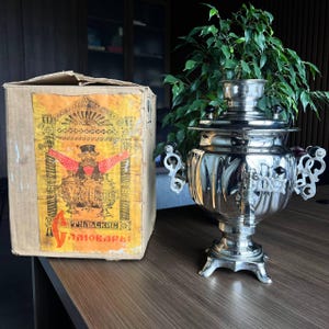 May include: A polished silver samovar with decorative handles and a tap is positioned beside a cardboard box with a vintage label. The box displays a colourful illustration of a man and samovars, with text in a foreign language. The samovar is on a wooden surface.