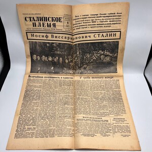 May include: An open newspaper with text in a foreign language, featuring a black and white photograph of a group of people and a deceased person. The headline reads "STALINSKOE PLEMYA" in bold letters. The paper is yellowed with age.