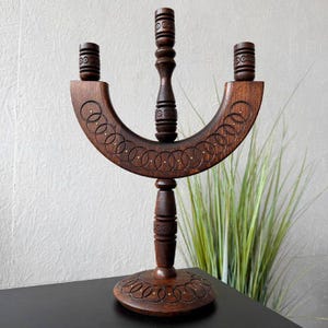 May include: A dark brown wooden candelabra featuring a curved horizontal bar with three candle holders. The wood is decorated with carved interlocking circle patterns and small inlaid metal dots along the base and the curved central section.