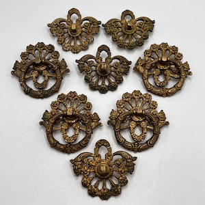 May include: Eight antique brass drawer pulls with ornate floral and ring designs. The pulls are in various shapes, including circular and fan-like, with a warm, aged patina. These decorative hardware pieces are ideal for vintage furniture restoration.