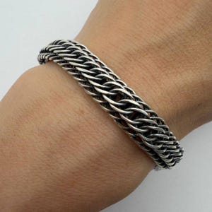 May include: A silver-colored bracelet with a woven chain design. The bracelet is made of multiple strands of metal, creating a textured and intricate pattern. The bracelet is worn on a wrist.