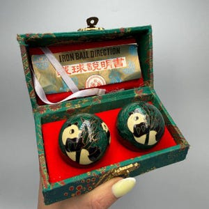 May include: A set of two green and black iron balls with panda designs, presented in a green box with red lining. The box has a gold clasp and a paper insert with text. The box is approximately 3 inches wide.