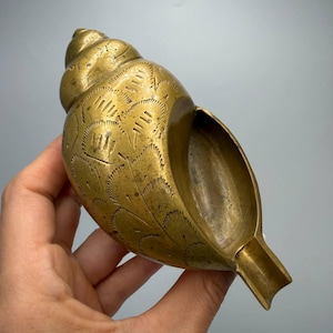 May include: A brass seashell-shaped object featuring an intricate, hand-engraved pattern of repeating arched lines across its surface. The metallic item has a smooth, hollow interior and a tapered, open end, showcasing a warm, golden-toned finish.