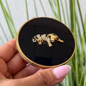 May include: A round, gold-toned compact mirror featuring a glossy black surface. Centered on the lid is a decorative, gold-toned tiger figurine embellished with small, clear, sparkling rhinestones along its body and head.