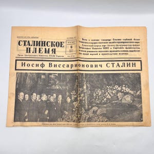 May include: An open newspaper with the headline "Stalin's Tribe" in Russian. The newspaper features a black and white photograph of a group of men and a deceased person. The text includes the name "Iosif Vissarionovich Stalin".