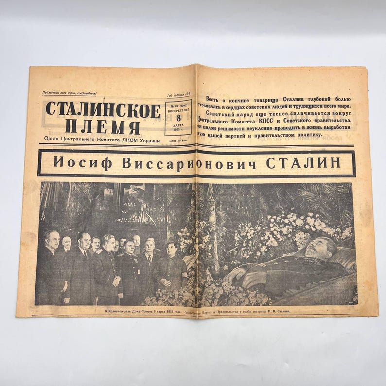 May include: An open newspaper with the headline "Stalin's Tribe" in Russian. The newspaper features a black and white photograph of a group of men and a deceased person. The text includes the name "Iosif Vissarionovich Stalin".