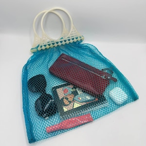 May include: A bright turquoise mesh tote bag with two white plastic circular handles. Inside the bag are a dark red rectangular wallet, a black-framed notebook with a graphic cover, black sunglasses, and a small pink cylindrical object.