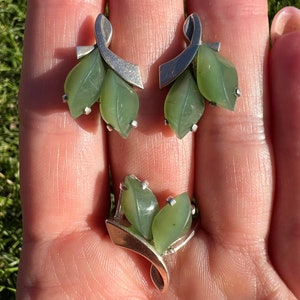 Op de afbeelding: Een set zilveren oorbellen en een ring met groene jade bladeren. De oorbellen hebben een gebogen zilveren ontwerp met drie jade bladeren elk. De ring heeft een vergelijkbaar ontwerp met twee jade bladeren.