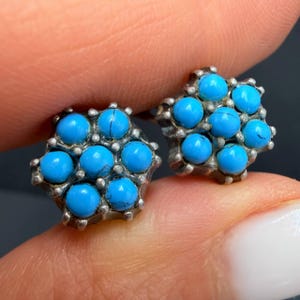 Pode incluir: Um par de brincos de tarracha com um grupo de sete pequenas pedras redondas de turquesa azul brilhante, cravadas numa armação de metal prateado. Cada brinco apresenta um padrão floral com uma pedra central rodeada por outras seis.