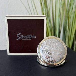 May include: A round silver-toned compact mirror featuring an intricate floral engraving on the lid, positioned in front of a square white and dark brown box. The box displays the text Stratton England Est. 1860 in a stylized silver script font.