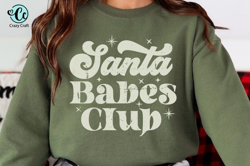 May include: Olive green sweatshirt with the text "Santa Babes Club" in a retro font. The text is in off-white with star accents. The Crazy Craft logo is visible in the upper left corner. The sweatshirt is a long-sleeved crew neck.