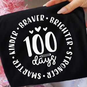 Op de afbeelding: Zwart T-shirt met een wit cirkelvormig ontwerp. Het ontwerp bevat de woorden "Kinder, Braver, Brighter, Stronger, Smarter" rond het getal "100 days" met een potloodafbeelding en hartjes. Het shirt wordt door een hand vastgehouden.
