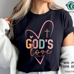 K&ouml;nnte beinhalten: Schwarzes T-Shirt mit einem herzf&ouml;rmigen Design und den Worten "GOD'S love" in bunten Buchstaben. Ein Kreuz befindet sich im Herzen, und der Bibelvers 1. Johannes 4,8 steht darunter. Das Crazy Craft-Logo befindet sich oben rechts.