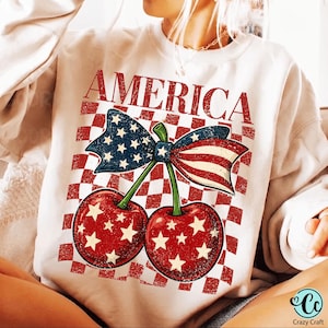Puede incluir: Una sudadera de cuello redondo color crema con un estampado gráfico de dos cerezas rojas con estrellas blancas, rematadas con un lazo con el patrón de la bandera estadounidense. El fondo es un cuadrado a cuadros rojos y blancos con la palabra AMERICA.