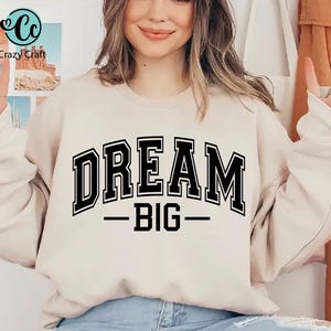 May include: A beige sweatshirt with the words "DREAM BIG" in bold, black, collegiate-style lettering. The text is centered and underlined. The Crazy Craft logo is visible in the upper left corner.