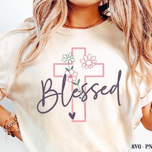 K&ouml;nnte beinhalten: Cremefarbenes T-Shirt mit einem rosa Kreuzdesign, floralen Akzenten und dem Wort "Blessed" in Schreibschrift. Ein kleines Herz befindet sich darunter. Das Shirt hat hochgekrempelte &Auml;rmel.