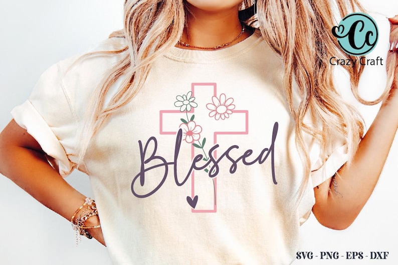 K&ouml;nnte beinhalten: Cremefarbenes T-Shirt mit einem rosa Kreuzdesign, floralen Akzenten und dem Wort "Blessed" in Schreibschrift. Ein kleines Herz befindet sich darunter. Das Shirt hat hochgekrempelte &Auml;rmel.