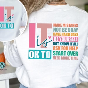 May include: White sweatshirt with a colorful graphic that says "It is OK to" and lists a series of phrases including "Make Mistakes", "Not Be Okay", "Have Hard Days", "Be Yourself", "Not Know It All", "Ask For Help", "Start Over", and "Need More Time".