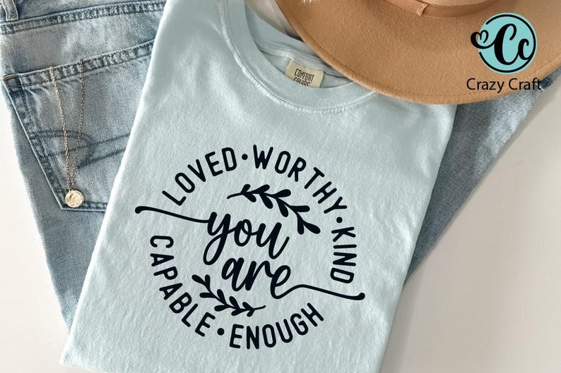 Pu&ograve; includere: Una maglietta blu chiaro con un design circolare nero che dice "Amato, Degno, Gentile, Capace, Abbastanza" con un design floreale e le parole "Tu Sei" al centro.