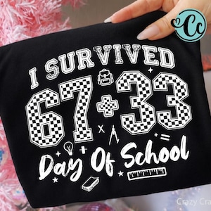 Puede incluir: Camiseta negra con el texto blanco "I Survived 67 + 33 Day Of School". Los números 67 y 33 tienen un patrón de cuadros. La camiseta también presenta gráficos escolares.