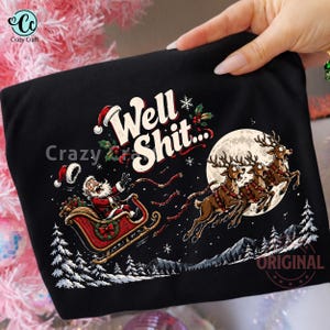 May include: A black t-shirt featuring a graphic of Santa Claus in a red sleigh pulled by three reindeer flying past a full moon. The text "Well Shit..." is printed in white above the sleigh. The scene includes snowy pine trees and snowflakes.