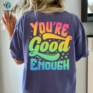 以下が含まれることがあります： 虹色の文字で「You're Good Enough」と書かれた紫色のTシャツです。
