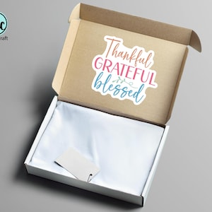 May include: An open cardboard box with a white interior and a sticker that reads "Thankful, Grateful, Blessed" in pink, blue, and coral. A blank tag is inside the box.