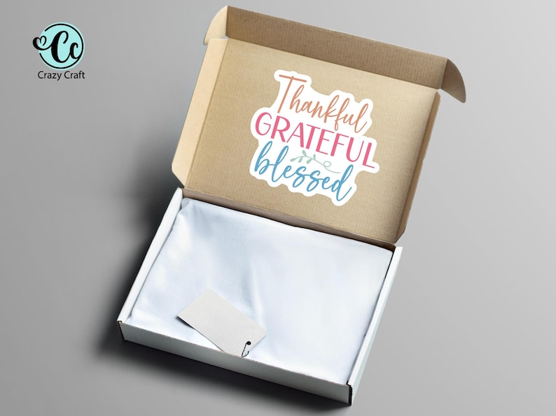May include: An open cardboard box with a white interior and a sticker that reads "Thankful, Grateful, Blessed" in pink, blue, and coral. A blank tag is inside the box.