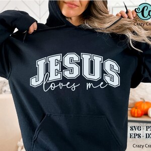 K&ouml;nnte beinhalten: Ein schwarzer Hoodie mit einer wei&szlig;en Grafik, auf der "JESUS loves me" in einer Schriftart im College-Stil steht.