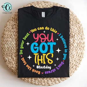 Puede incluir: Camiseta negra con un diseño circular de colores que dice "You Got This" con el texto "Do your best", "You can do this", "I believe in you", "Don't stress", "Rock the test" y "#testday" alrededor del círculo.