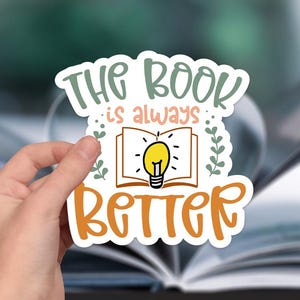 May include: A white sticker with the text "The book is always better" in orange and pink lettering. A lightbulb is inside an open book, with green leaves on either side.