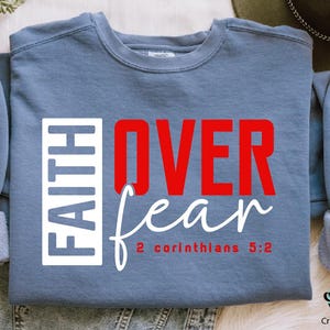 May include: A blue sweatshirt with the words "FAITH OVER fear" in red and white lettering. The text "2 Corinthians 5:2" is printed in white below the main text.