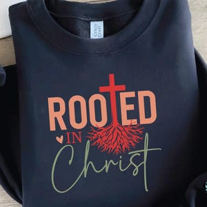 K&ouml;nnte beinhalten: Ein schwarzes Sweatshirt mit dem Schriftzug "ROOTED IN Christ" in Koralle und Gr&uuml;n. Ein rotes Kreuz und ein Wurzeldesign sind &uuml;ber dem Text zentriert. Das Sweatshirt hat einen Rundhalsausschnitt und lange &Auml;rmel.