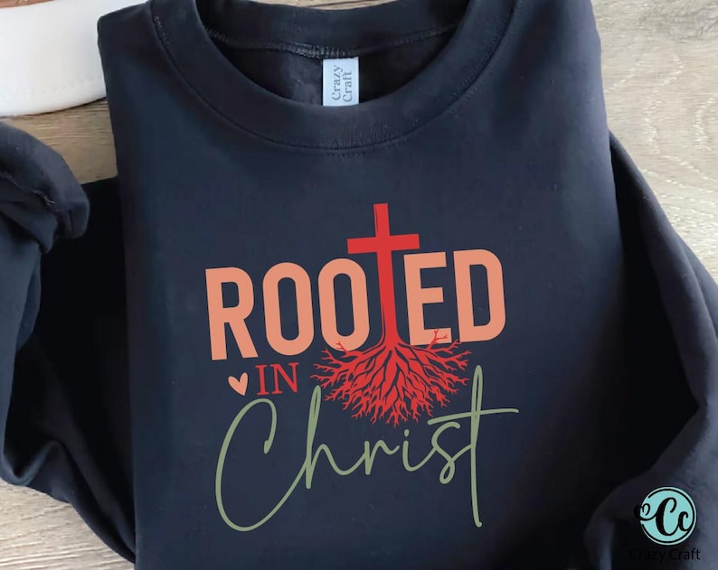 K&ouml;nnte beinhalten: Ein schwarzes Sweatshirt mit dem Schriftzug "ROOTED IN Christ" in Koralle und Gr&uuml;n. Ein rotes Kreuz und ein Wurzeldesign sind &uuml;ber dem Text zentriert. Das Sweatshirt hat einen Rundhalsausschnitt und lange &Auml;rmel.