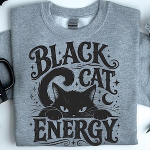 May include: A heather gray crewneck sweatshirt featuring a black graphic print of a cat with the text Black Cat Energy. Beside the sweatshirt is a black mug with the white text Vampire Juice. The design includes small star and moon accents.