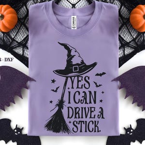 K&ouml;nnte beinhalten: Ein helllila T-Shirt mit einem Hexenhut- und Besen-Motiv. Der Text lautet "YES I CAN DRIVE A STICK." Das Design umfasst Flederm&auml;use und Sterne. Dekorative K&uuml;rbisse und Flederm&auml;use umgeben das Shirt, perfekt f&uuml;r Halloween.