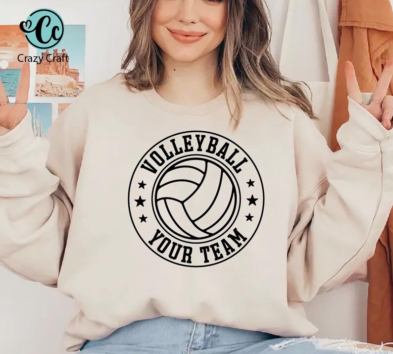 K&ouml;nnte beinhalten: Ein beiges Sweatshirt mit einem schwarz-wei&szlig;en Volleyball-Design. Das Design zeigt eine Volleyball-Grafik in einem kreisf&ouml;rmigen Rahmen mit den Worten "VOLLEYBALL" und "YOUR TEAM" in fetter Schrift. Das Sweatshirt ist l&auml;ssige Sportbekleidung.