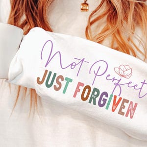 K&ouml;nnte beinhalten: Wei&szlig;es Sweatshirt mit dem Text "Not Perfect Just Forgiven" in verschiedenen Farben. &Uuml;ber dem Wort "Perfect" befindet sich eine kleine Blumengrafik. Eine Person h&auml;lt das Sweatshirt und eine wei&szlig;e Tasse.