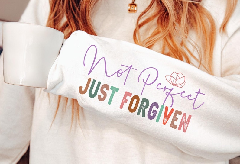 K&ouml;nnte beinhalten: Wei&szlig;es Sweatshirt mit dem Text "Not Perfect Just Forgiven" in verschiedenen Farben. &Uuml;ber dem Wort "Perfect" befindet sich eine kleine Blumengrafik. Eine Person h&auml;lt das Sweatshirt und eine wei&szlig;e Tasse.