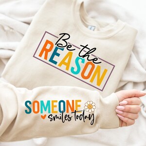 以下が含まれることがあります： ベージュのトレーナーに、カラフルな枠の中に「Be the REASON」という文字と、袖にスマイリーフェイス付きの「SOMEONE smiles today」という文字が書かれています。