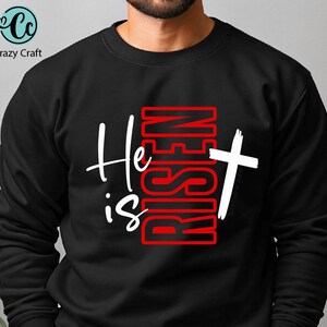 May include: Black sweatshirt with the text "He is Risen" in white and red. The design includes a white cross. The sweatshirt is from Crazy Craft.