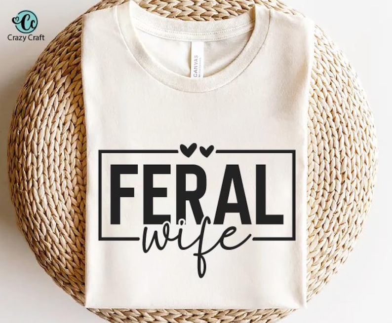 May include: White t-shirt with black text that reads "Feral Wife" with two small hearts above the text.