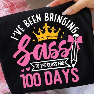 以下が含まれることがあります： 「I've Been Bringing Sass to the Class for 100 Days」の文字が白とピンクでプリントされた黒いTシャツ。デザインには金の王冠、ピンクのリボン、鉛筆が含まれています。Tシャツは手で持たれています。