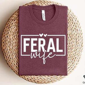 May include: A maroon t-shirt with a white graphic that says "Feral Wife" with two small hearts above the text.