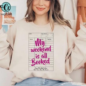 May include: A beige sweatshirt with the text "My weekend is all Booked" in a pink font, designed to look like a library card. The sweatshirt also has the Crazy Craft logo in the upper left corner.