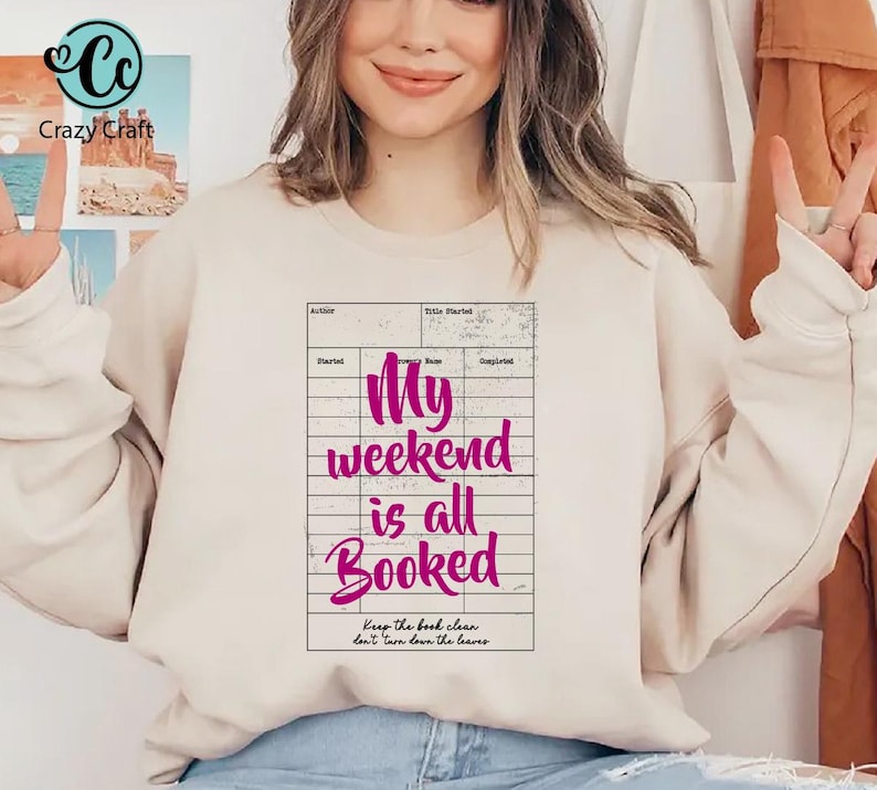 May include: A beige sweatshirt with the text "My weekend is all Booked" in a pink font, designed to look like a library card. The sweatshirt also has the Crazy Craft logo in the upper left corner.