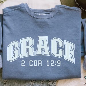 K&ouml;nnte beinhalten: Ein blaues Sweatshirt mit dem Wort "GRACE" in wei&szlig;en umrandeten Buchstaben mit einem wei&szlig;en Umriss. Der Text "2 COR 12:9" ist unter dem Wort "GRACE" gedruckt.
