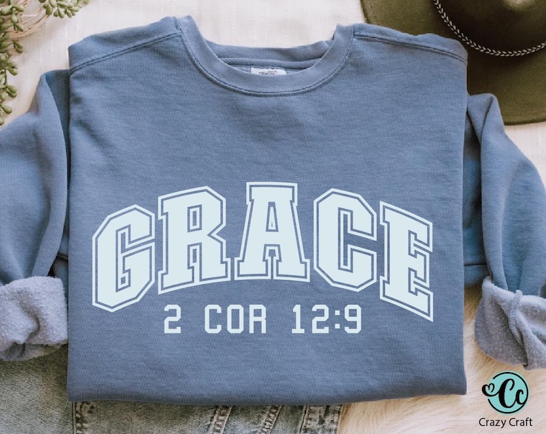 K&ouml;nnte beinhalten: Ein blaues Sweatshirt mit dem Wort "GRACE" in wei&szlig;en umrandeten Buchstaben mit einem wei&szlig;en Umriss. Der Text "2 COR 12:9" ist unter dem Wort "GRACE" gedruckt.